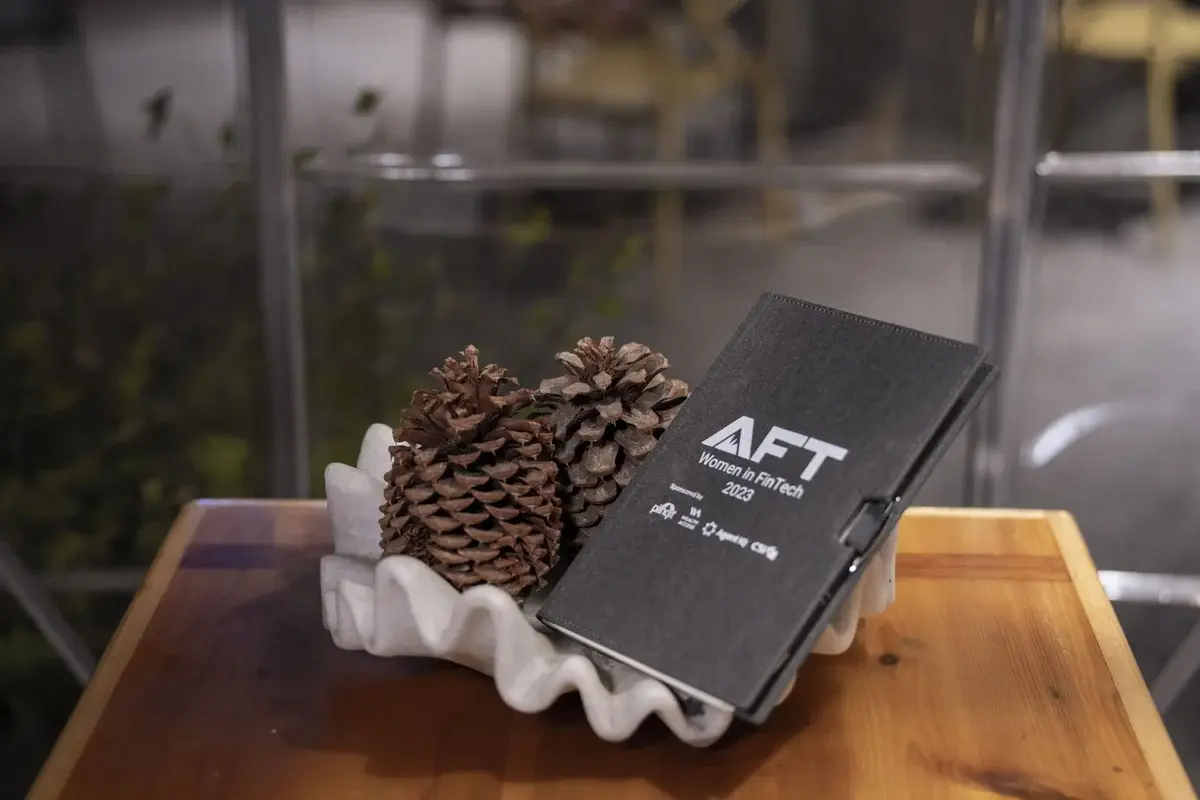 AFT Women in FinTech 2023 — branded notebook with AFT Women in FinTech 2023 cover displayed alongside pine cones